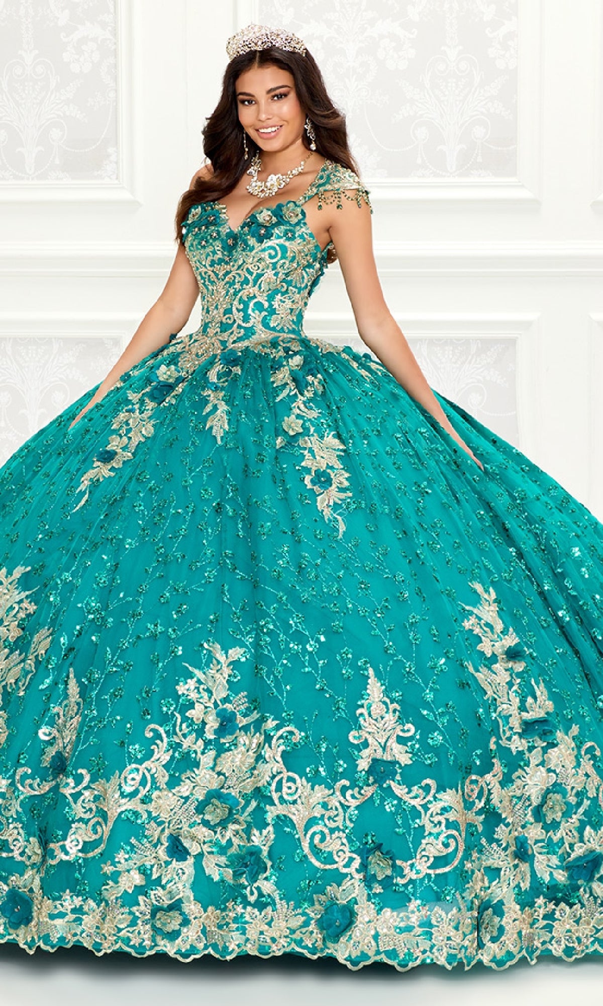 A woman in the Quinceanera Dress PR30088 By Princesa, a turquoise gown with gold embroidery and lace details, wears a tiara and stands before a white ornate wall.